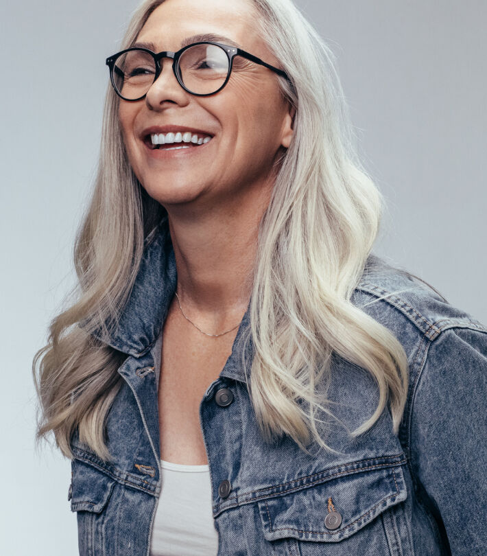 Joyful woman with long white hair and glasses with lens smiling warmly against a light gray background