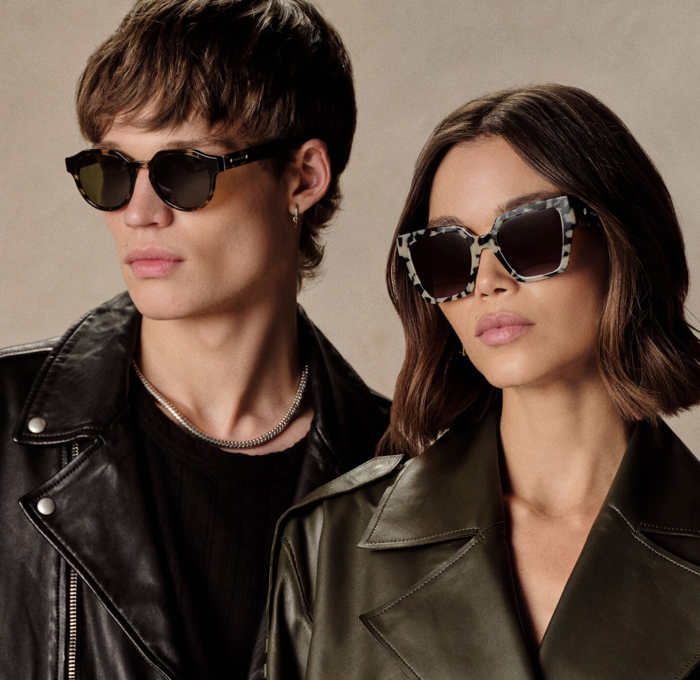Man in leather jacket and woman in dark coat wearing AllSaints sunglasses, exuding cool confidence.