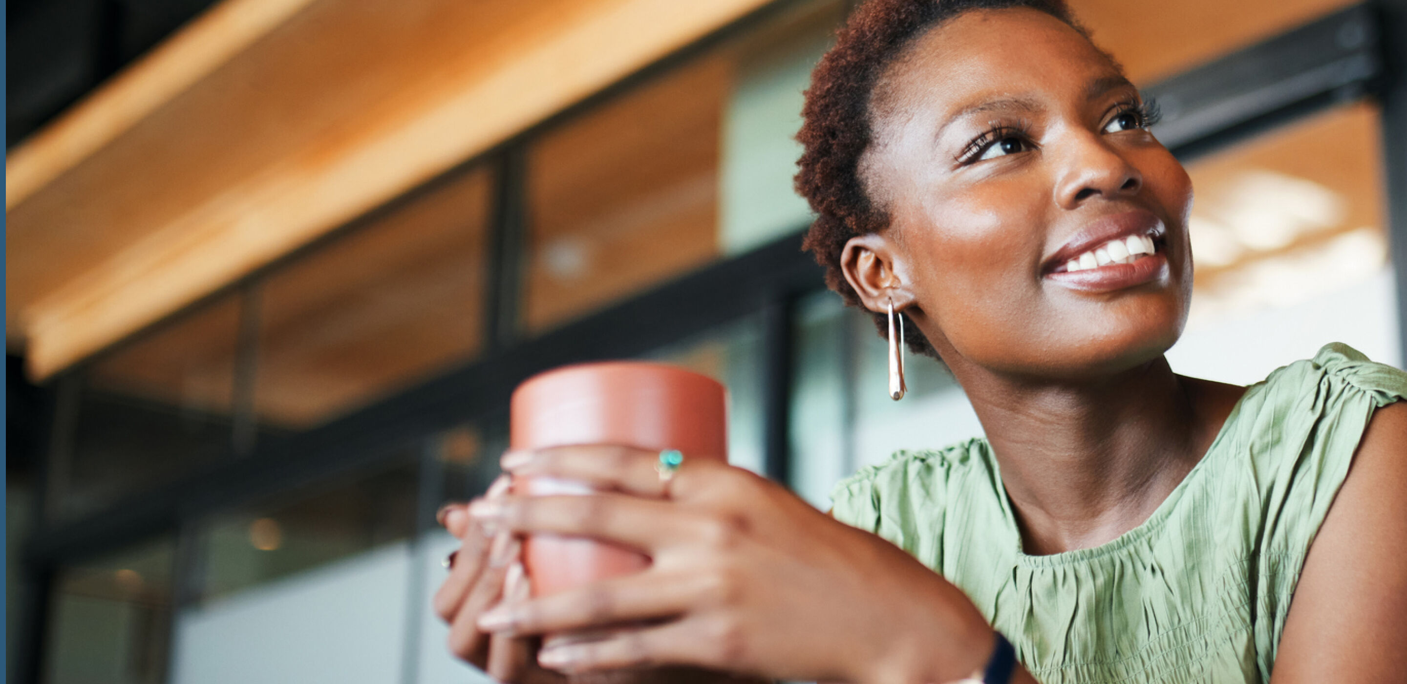 Smiling woman with short hair wearing a green top and holding a red cup in a softly lit room.
