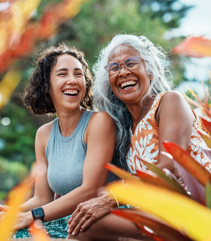 Two women sitting outdoors laughing together, surrounded by greenery and colourful foliage.