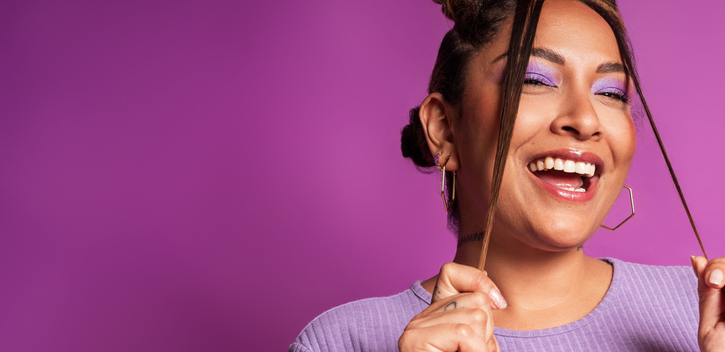 Smiling woman wearing purple eyeshadow and a lavender top, representing makeup suitable for contact lens wearers.