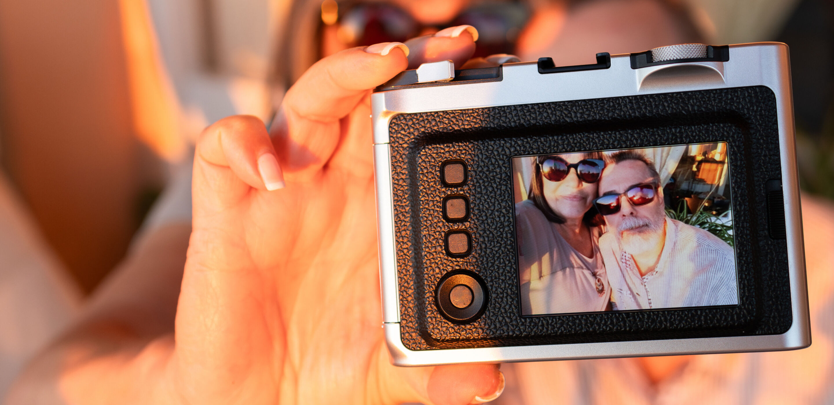 A hand holds a camera, displaying a screen showing a smiling man and woman in sunglasses.