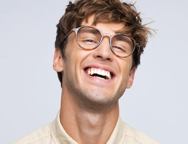 A smiling man wearing brown Boots Opticians glasses and a light beige shirt against a plain background