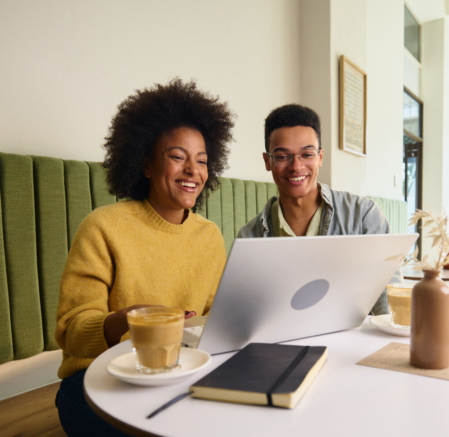 A smiling woman in a yellow sweater and a man wearing glasses with Acuvue lens sit at a caf&eacute; table with a laptop.