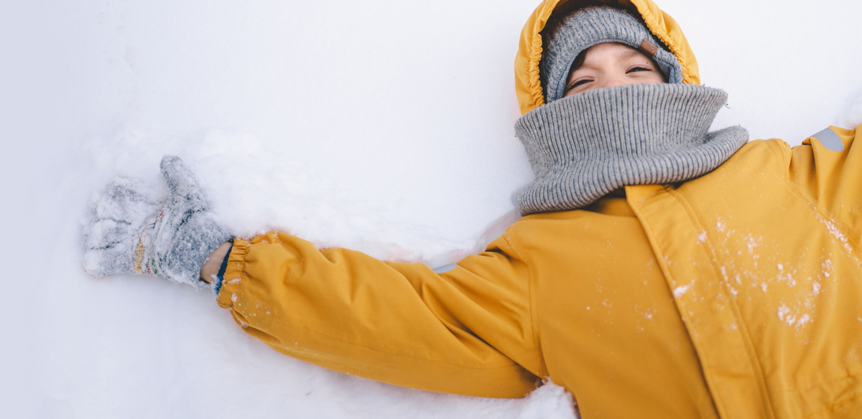 Person in a yellow jacket and gray scarf lies on snow, smiling with arms spread wide. 