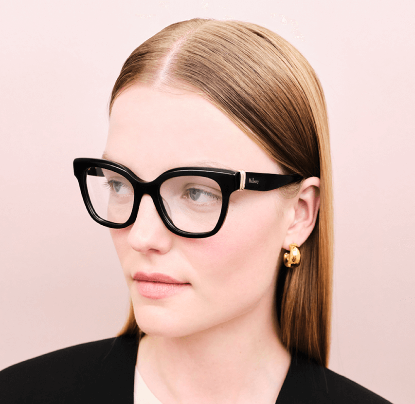 Woman wearing Mulberry black glasses with gold hoop earrings against a soft pink background