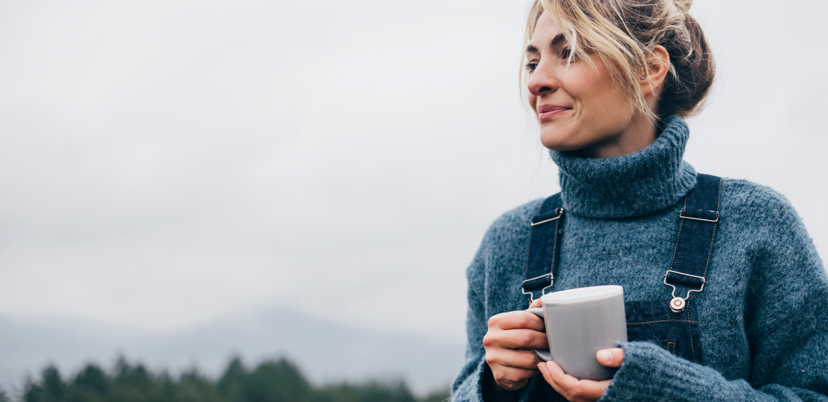 Woman in blue sweater holding a mug outdoors under overcast sky, conveying calm and reflection