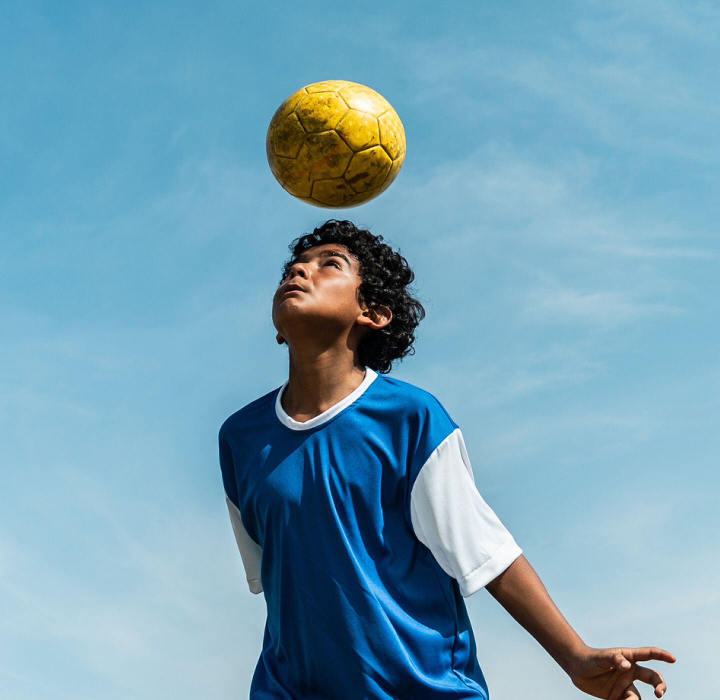 Boy playing football wearing MiSight 1 day contact lenses for myopia control, focusing on a ball against a blue sky