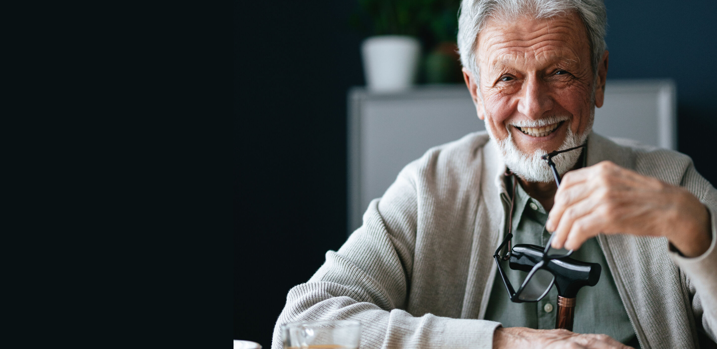 Smiling elderly man with grey hair and beard holding glasses in a softly lit setting.