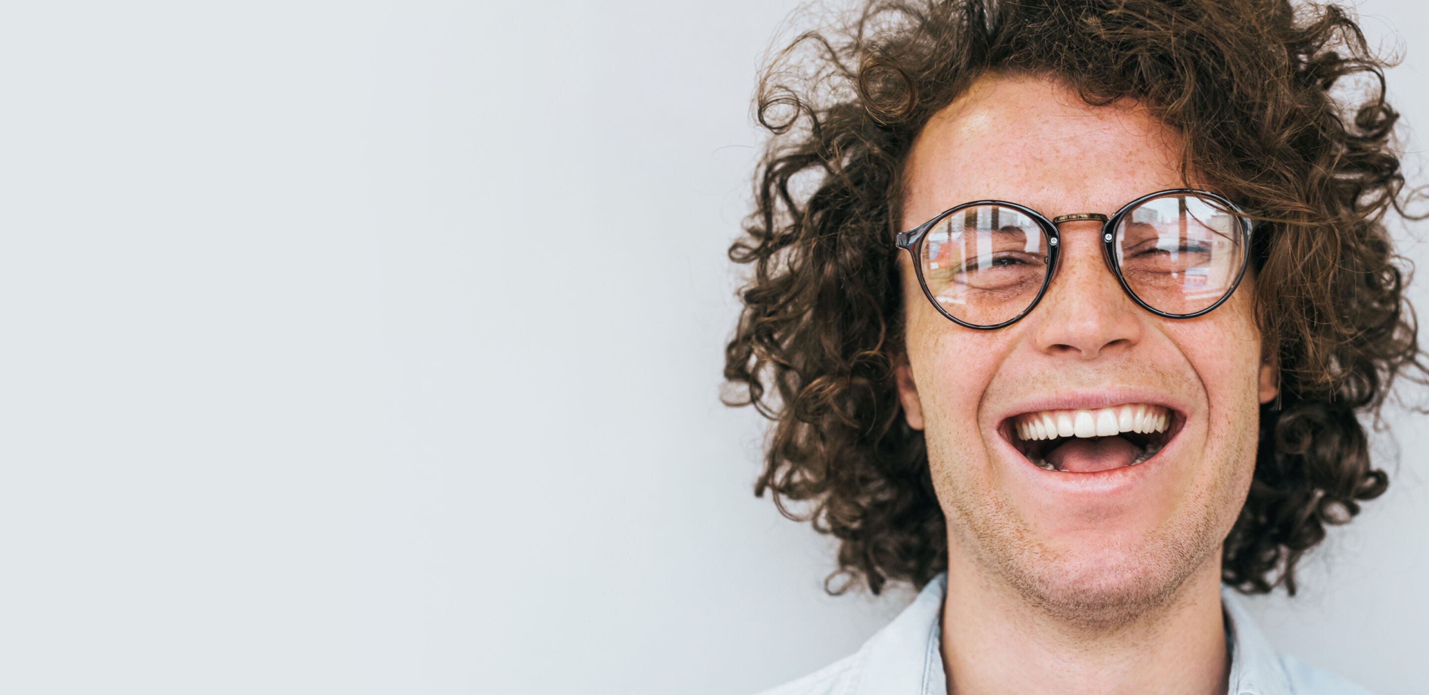 Young person with curly hair and glasses smiling broadly against a plain background