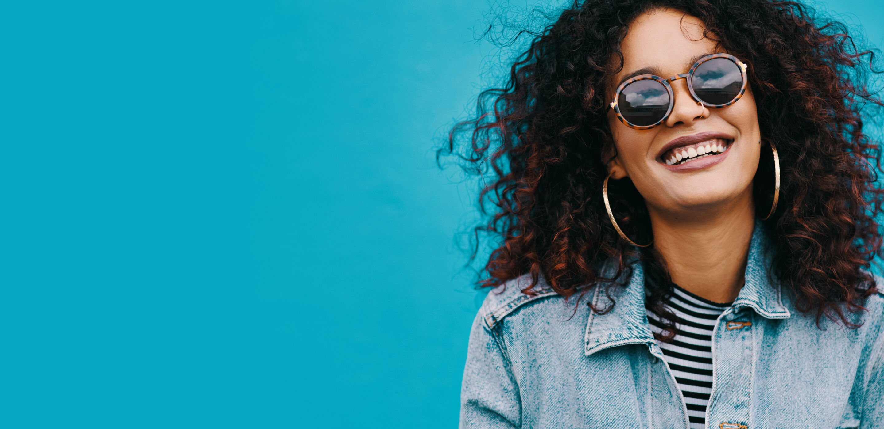 A woman with curly hair wearing round Boots Opticians sunglasses, smiling against a bright blue background