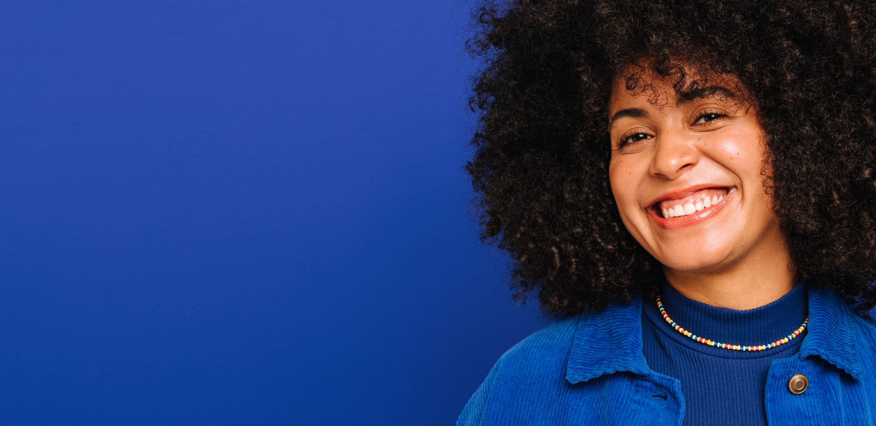 Smiling women with curly hair in a blue jacket and beaded necklace against blue background