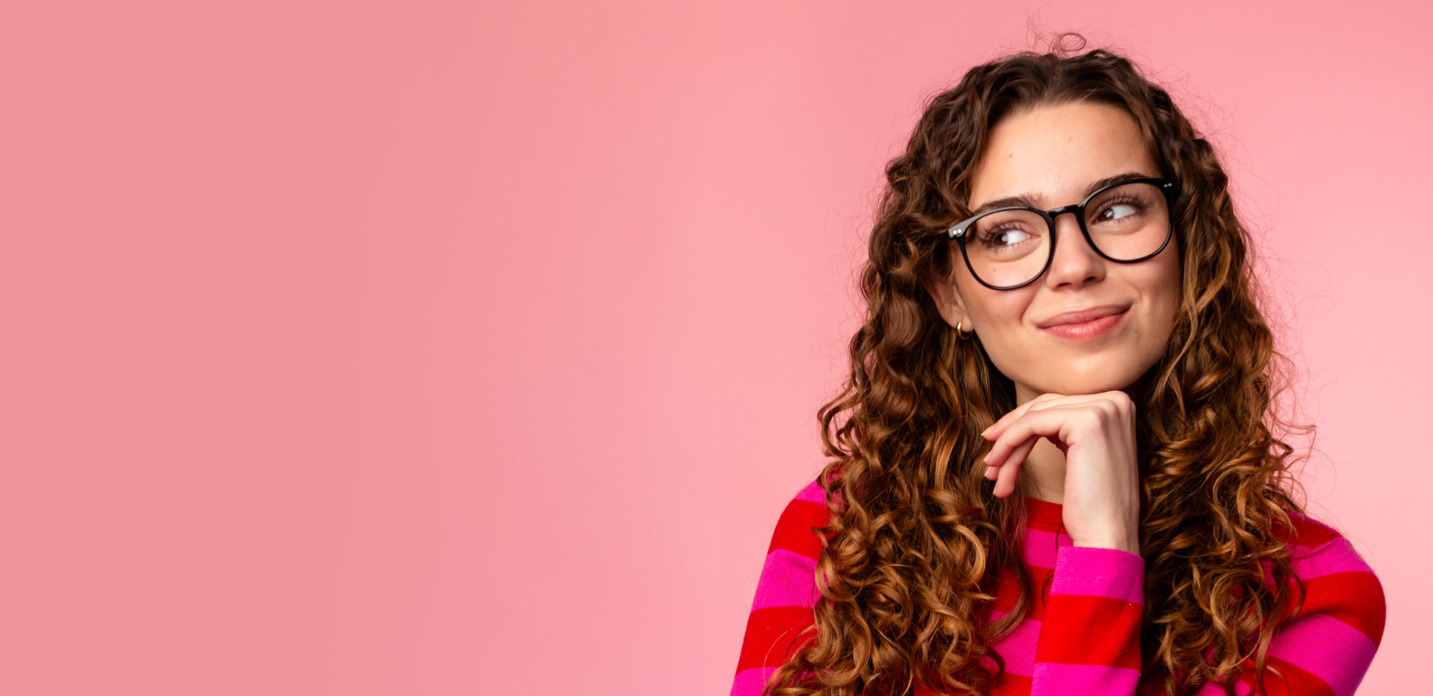 Young woman with curly hair wearing glasses and a striped sweater, smiling against a pink background.