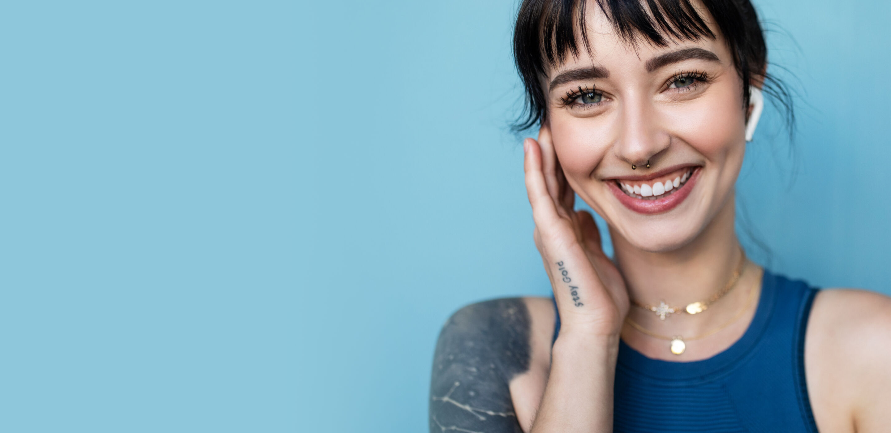 Smiling woman with bangs wearing earbuds and a sleeveless top against a blue background.