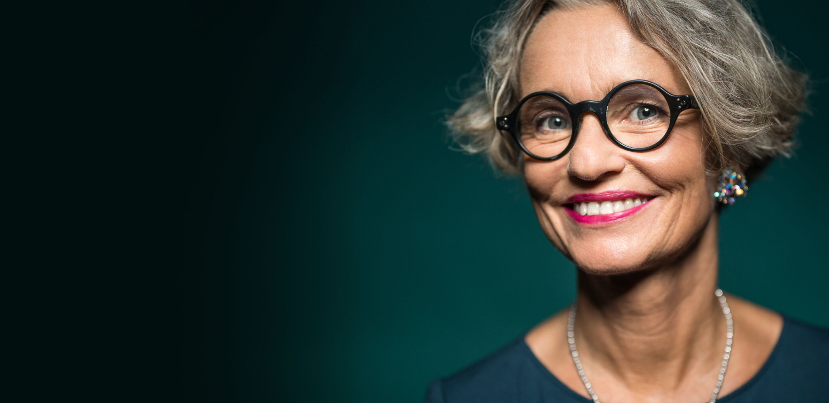 Smiling older woman with short grey hair wearing round black glasses and colourful earrings against a dark teal background.