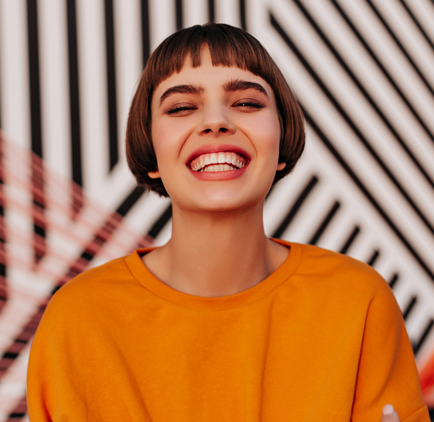 Smiling girl with a bob haircut wearing an orange top against a black and white striped background.