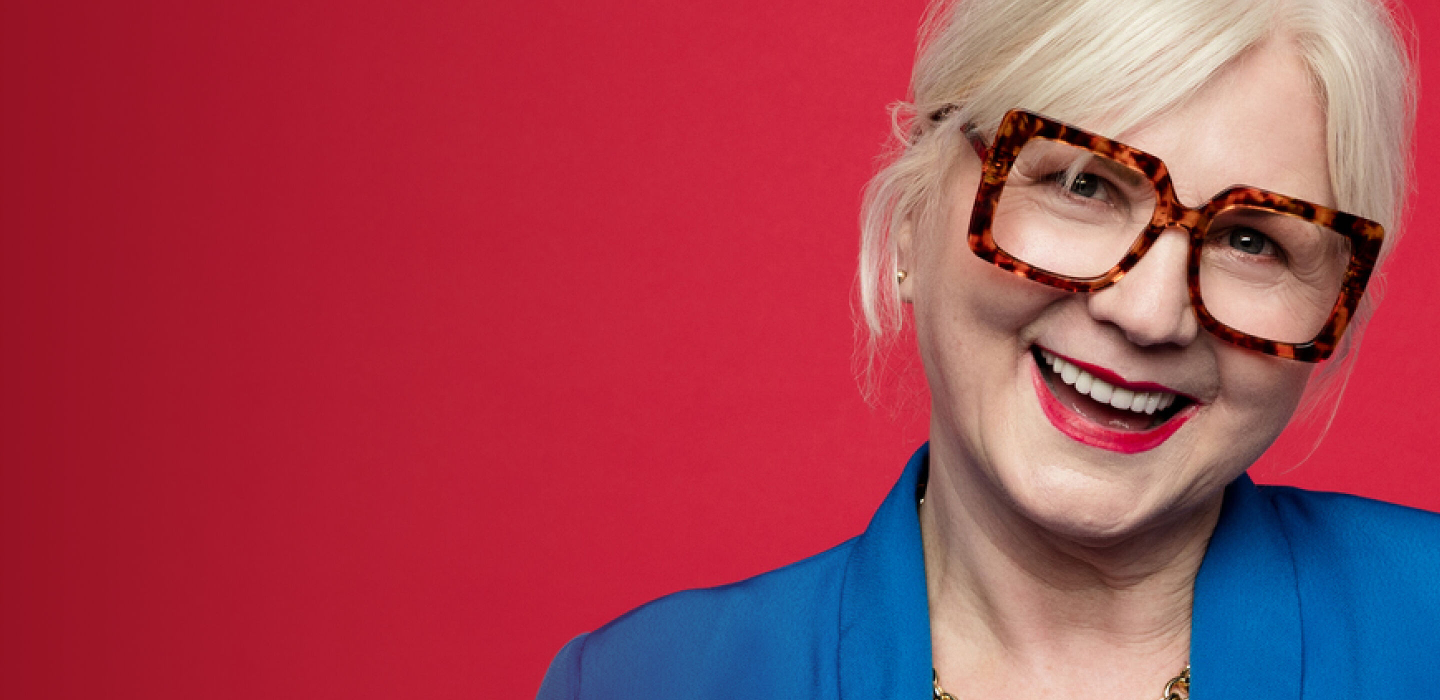 An elderly woman wearing tortoiseshell Boots Opticians glasses, smiling against a vibrant red background