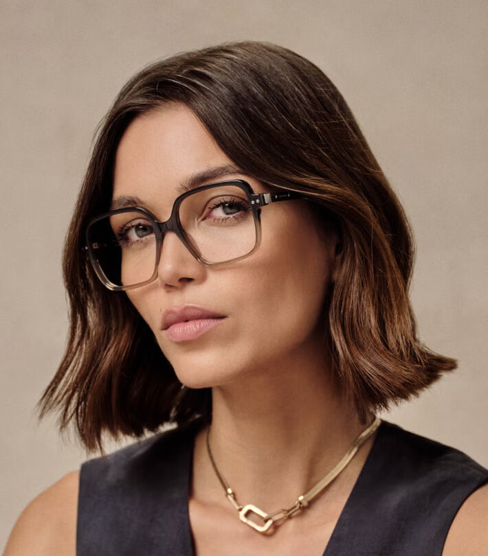 Woman with black AllSaints glasses, dark sleeveless top, and gold necklace gazing at camera on beige background