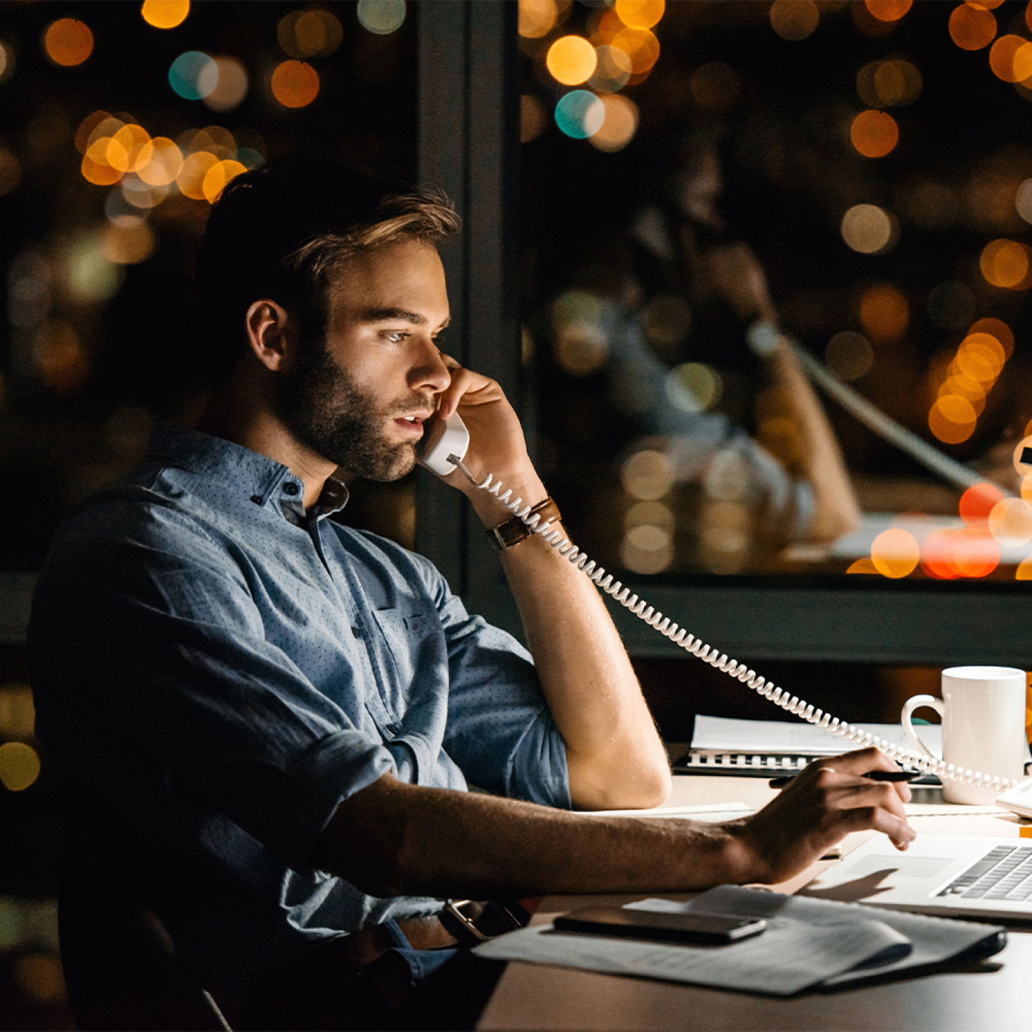 Man working late at a desk, talking on the phone with city lights blurred in the background