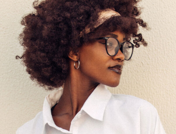 Woman with curly hair wearing boots opticians&rsquo; glasses and a white collared shirt, looking to the side.