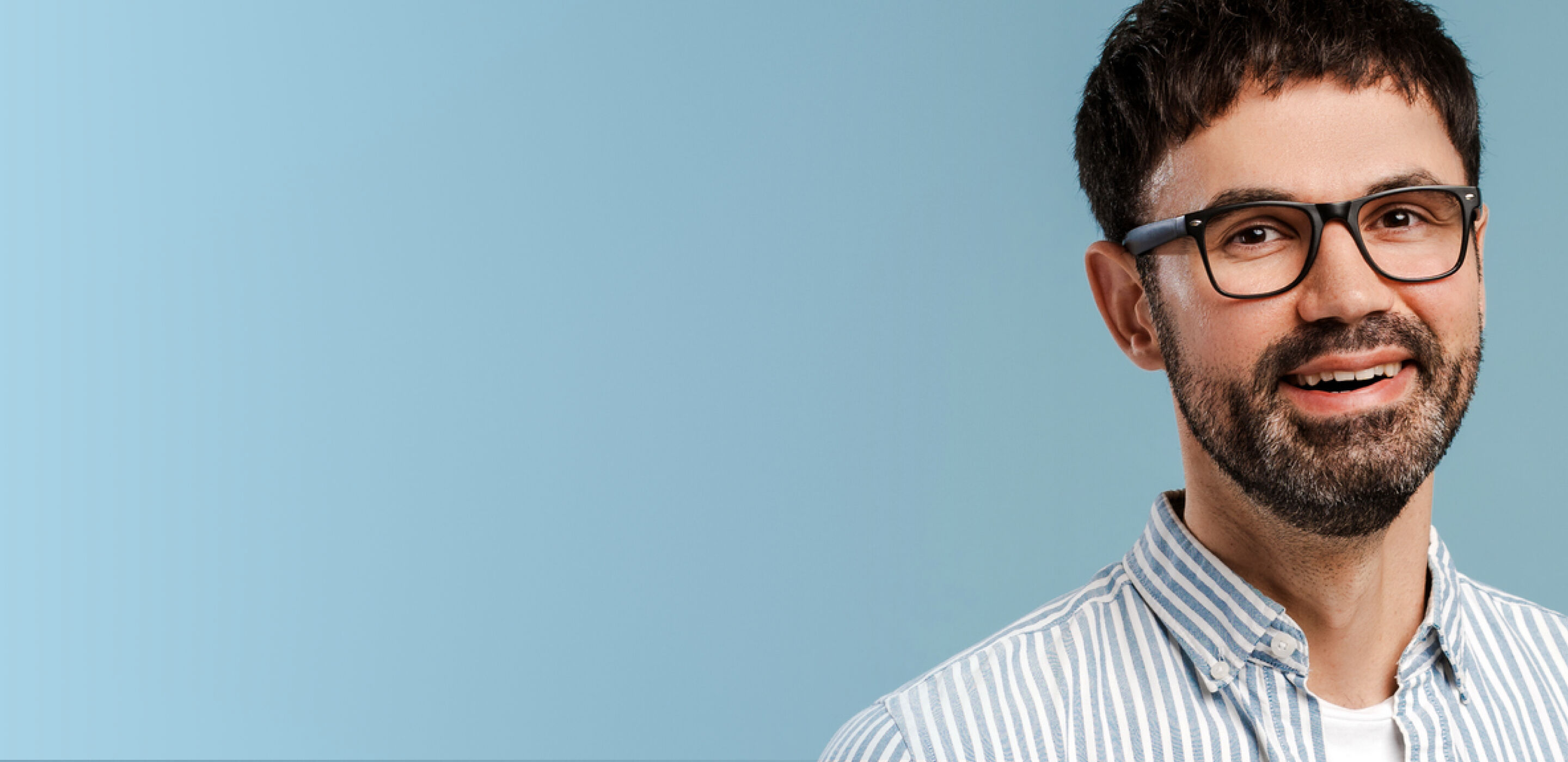 Smiling man with beard and boots optician&rsquo;s glasses in a striped shirt against a soft blue background.