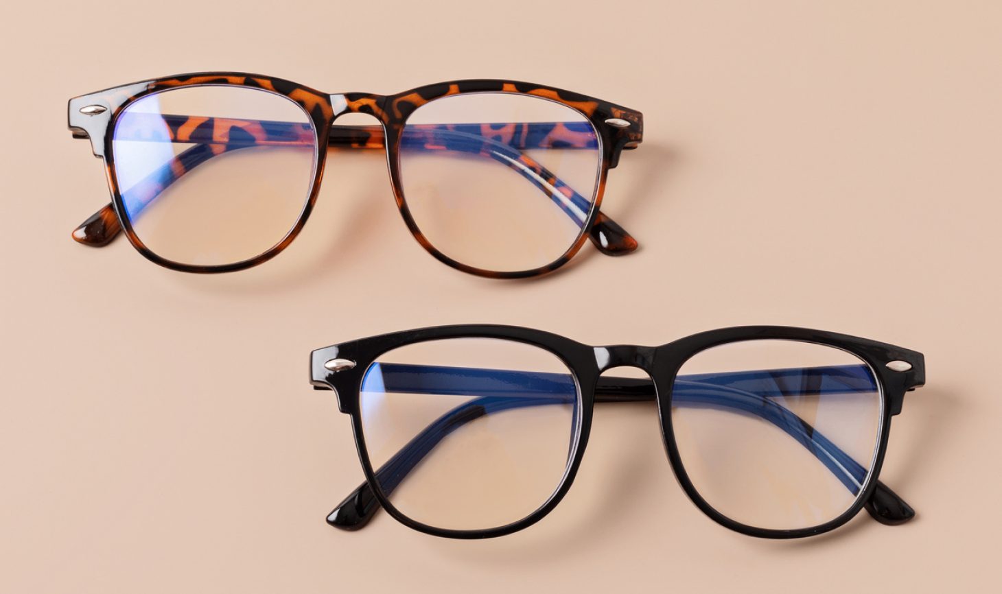 Two pairs of boots opticians eyeglasses on a beige background. The top pair has tortoiseshell frames, and the bottom pair has black frames. Both have clear lenses.