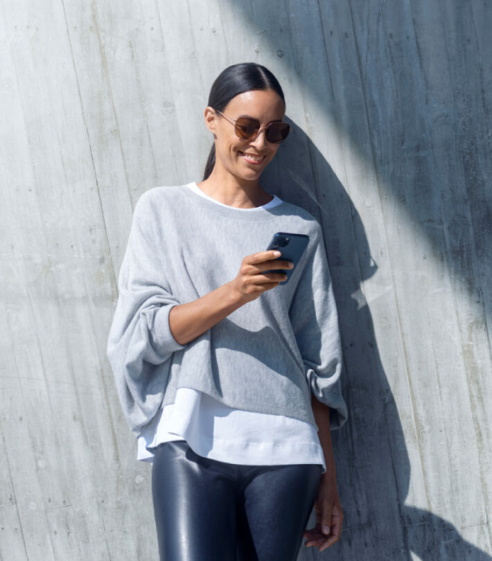 Woman wearing sunglasses with Zeiss PhotoFusion X lens smiling at her phone while leaning against a concrete wall in sunlight.