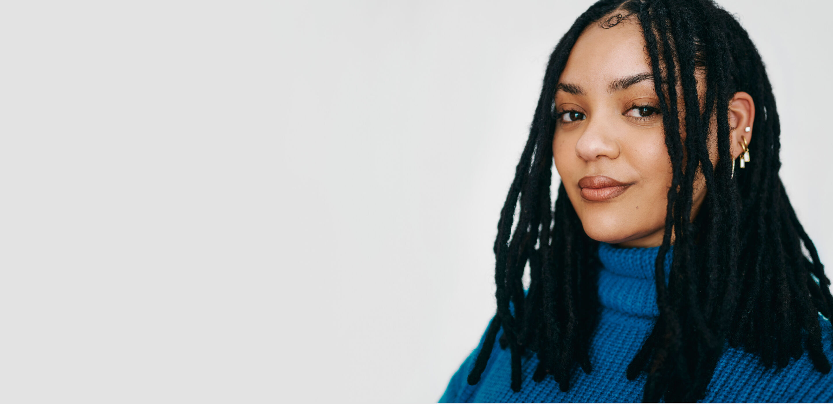 Smiling woman with long black dreadlocks wearing a blue turtleneck against a white background.