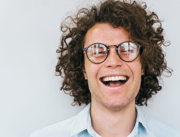 Young person with curly hair and glasses smiling broadly against a plain background