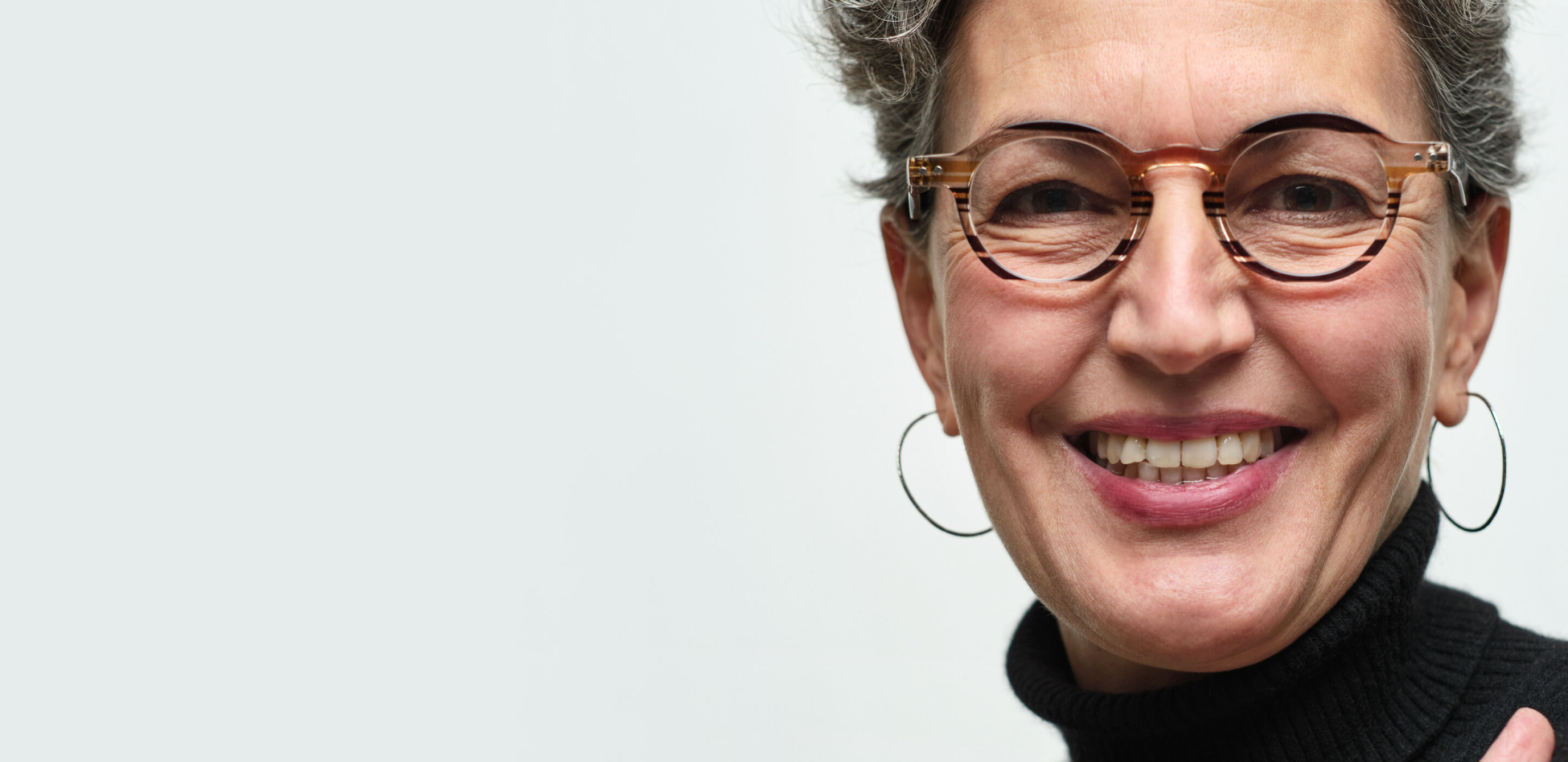 Smiling woman with short gray hair wearing round tortoiseshell glasses with varifocal lens and hoop earrings..