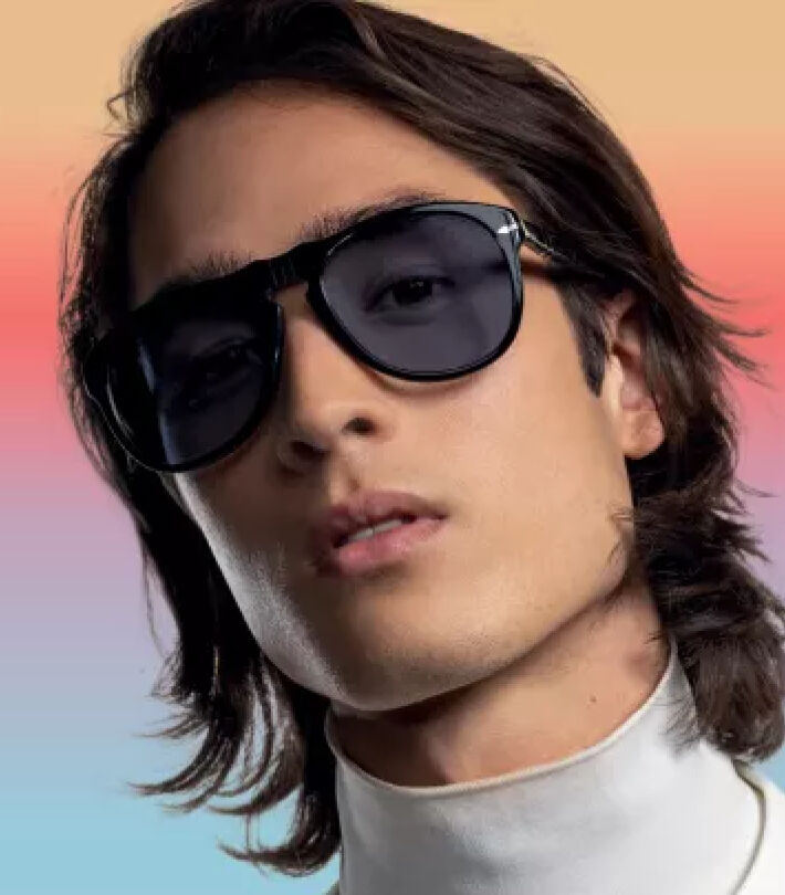 Man with long hair wearing large dark sunglasses with transtitions lenses and a white turtleneck against a soft gradient background.