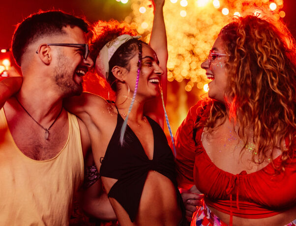 Three friends smiling together at a summer festival, wearing sunglasses under colourful lights